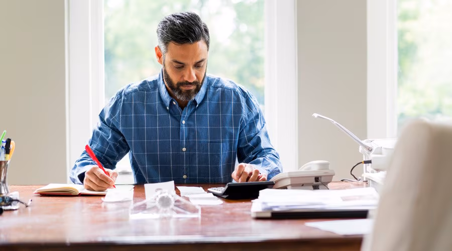 A man sits at a desk using a calculator and taking notes, working in a bright home office.